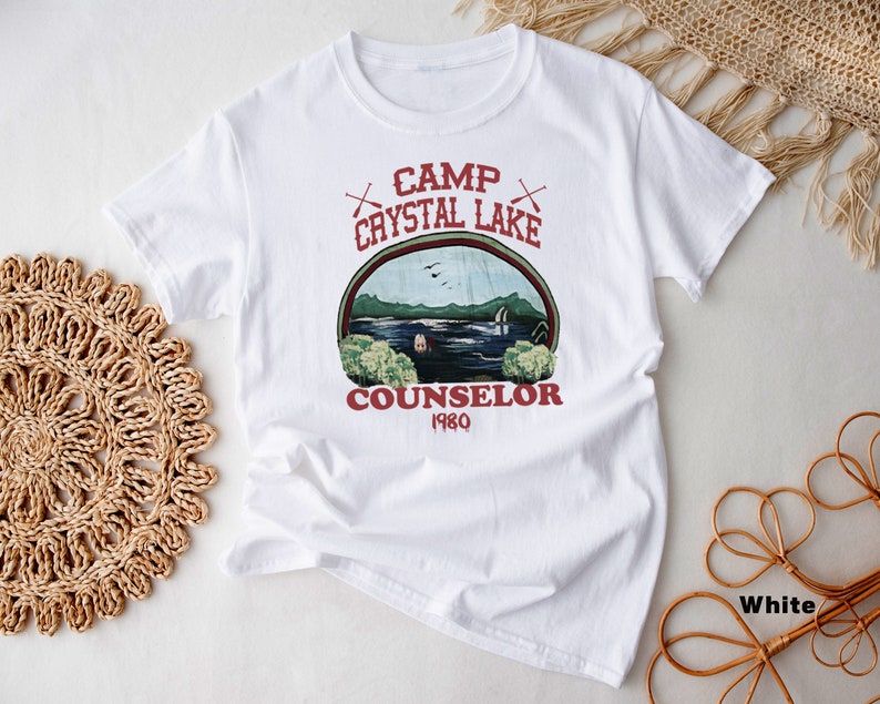May include: White t-shirt with a graphic featuring the text "Camp Crystal Lake Counselor 1980" in red. The graphic depicts a lake scene with trees, a cabin, and the text "Camp Crystal Lake" in an arch. The shirt is made of a soft material.