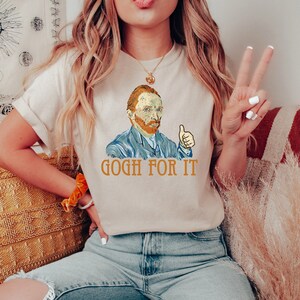 May include: A beige t-shirt with a graphic of Vincent van Gogh giving a thumbs up and the text "GOGH FOR IT".