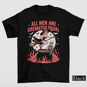 May include: Black t-shirt with a Halloween-themed graphic. The design features a witch on a broom, a skeleton in a cauldron, and the text "ALL MEN ARE CREMATED EQUAL" in pink and white. The image includes bats and flames.