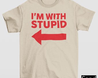 May include: Sand-colored t-shirt with the red text "I'M WITH STUPID" and a red arrow pointing left. The shirt is a crew neck style.