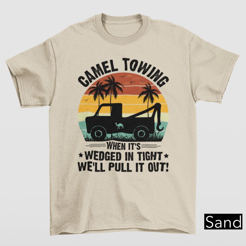 May include: A tan t-shirt with a black graphic design. The design features a tow truck with a camel in the bed of the truck. The tow truck is driving on a beach with palm trees and a sunset in the background. The text on the graphic reads 'Camel Towing When It's Wedged In Tight We'll Pull It Out!'.