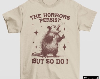 Könnte beinhalten: Ein beiges T-Shirt mit einer braunen Opossum-Illustration und dem Text "The Horrors Persist But So Do I".