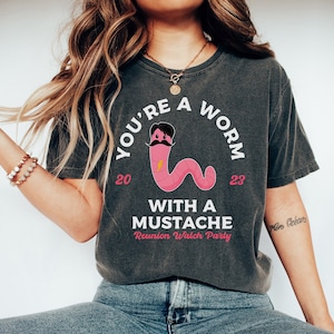 May include: A dark gray t-shirt with a white graphic of a pink worm with a black mustache and the text "You're a worm 20 23 with a mustache Reunion Watch Party".