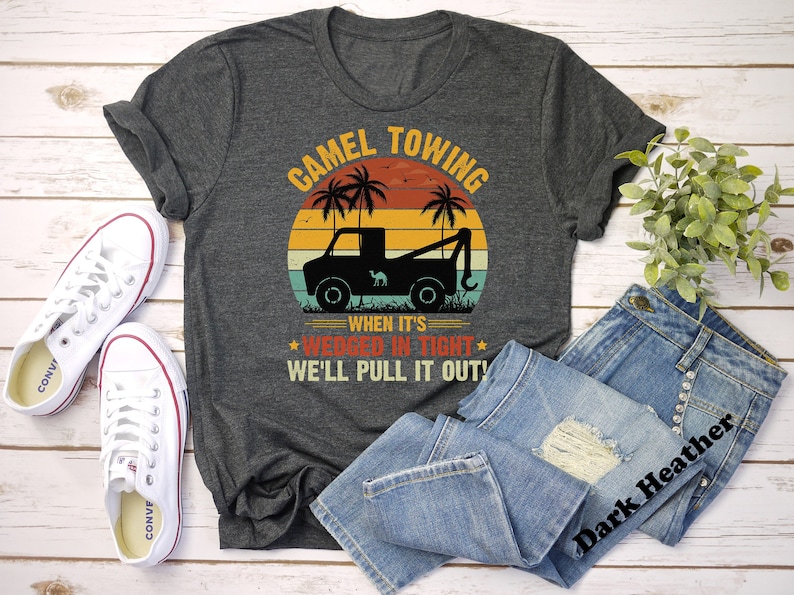 May include: A dark heather grey t-shirt with a vintage-style graphic of a tow truck pulling a camel. The graphic includes palm trees and a sunset. The text on the shirt reads "Camel Towing When It's Wedged In Tight We'll Pull It Out!"