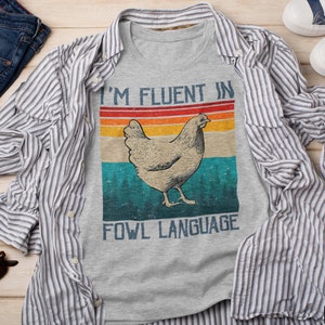 May include: A grey t-shirt with a retro-style graphic of a chicken and the text "I'm fluent in fowl language". The shirt is on a wooden surface with a blue and white striped shirt, blue jeans, and sunglasses.