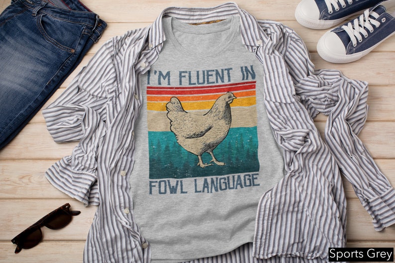 May include: A grey t-shirt with a retro-style graphic of a chicken and the text "I'm fluent in fowl language". The shirt is on a wooden surface with a blue and white striped shirt, blue jeans, and sunglasses.