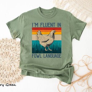 May include: A green t-shirt with a retro-style graphic of a chicken and the text "I'm fluent in fowl language".