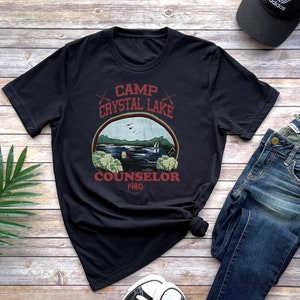 May include: Black t-shirt with a graphic featuring a lake scene and the text "Camp Crystal Lake Counselor 1980." The design includes a lake, trees, and birds. The shirt is paired with jeans and a black cap.