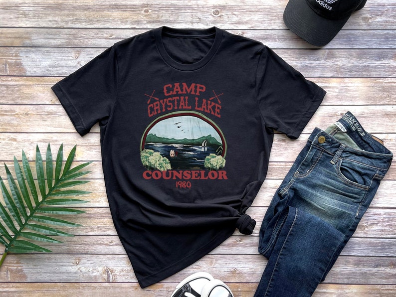 May include: Black t-shirt with a graphic featuring a lake scene and the text "Camp Crystal Lake Counselor 1980." The design includes a lake, trees, and birds. The shirt is paired with jeans and a black cap.
