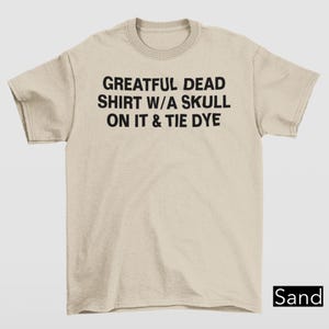 May include: Sand-colored t-shirt with the text "Grateful Dead Shirt W/A Skull On It & Tie Dye" in black. The shirt has short sleeves and a crew neck.