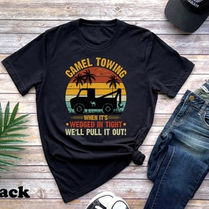 May include: Black t-shirt with a vintage-style graphic of a tow truck pulling a camel. The text reads "Camel Towing When It's Wedged In Tight We'll Pull It Out!"