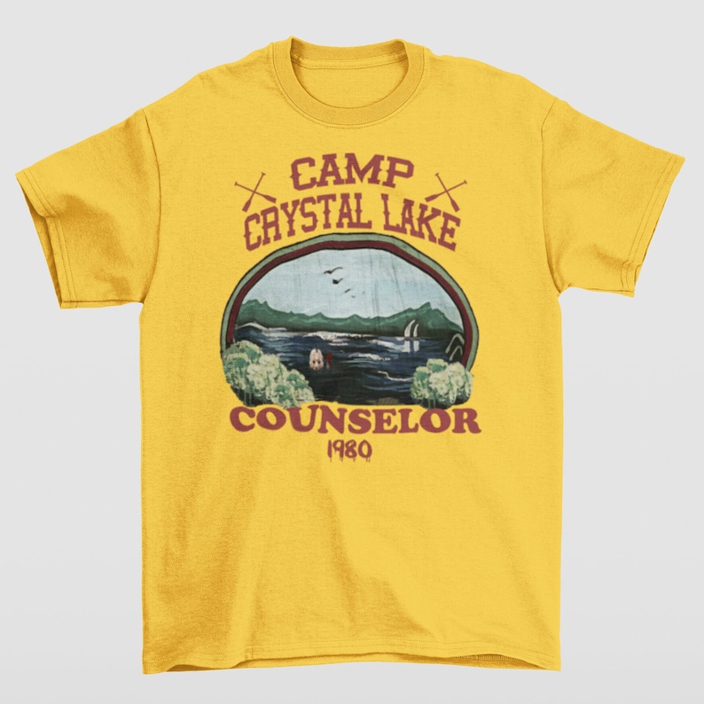 May include: A yellow t-shirt featuring a graphic design with the text "CAMP CRYSTAL LAKE COUNSELOR 1980". The design includes an oval image of a lake scene with trees, water, and a cabin. Two crossed axes are above the text.