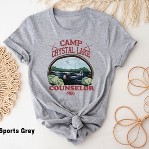 May include: Sports grey t-shirt featuring a graphic design with the text "Camp Crystal Lake Counselor 1980". The design includes a lake scene with trees, a cabin, and the text in a circular frame. The shirt is knotted at the bottom.