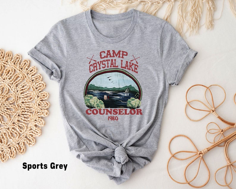 May include: Sports grey t-shirt featuring a graphic design with the text "Camp Crystal Lake Counselor 1980". The design includes a lake scene with trees, a cabin, and the text in a circular frame. The shirt is knotted at the bottom.