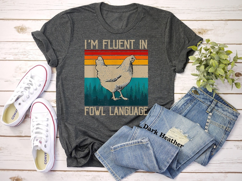 May include: A dark heather grey t-shirt with a vintage-style graphic of a chicken and the text "I'm fluent in fowl language".
