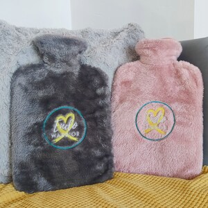 Op de afbeelding: Twee pluche warmwaterkruiken met geborduurde gele linten en de tekst "Endo Warrior" in een cirkel. Een kruik is grijs en de andere is roze.