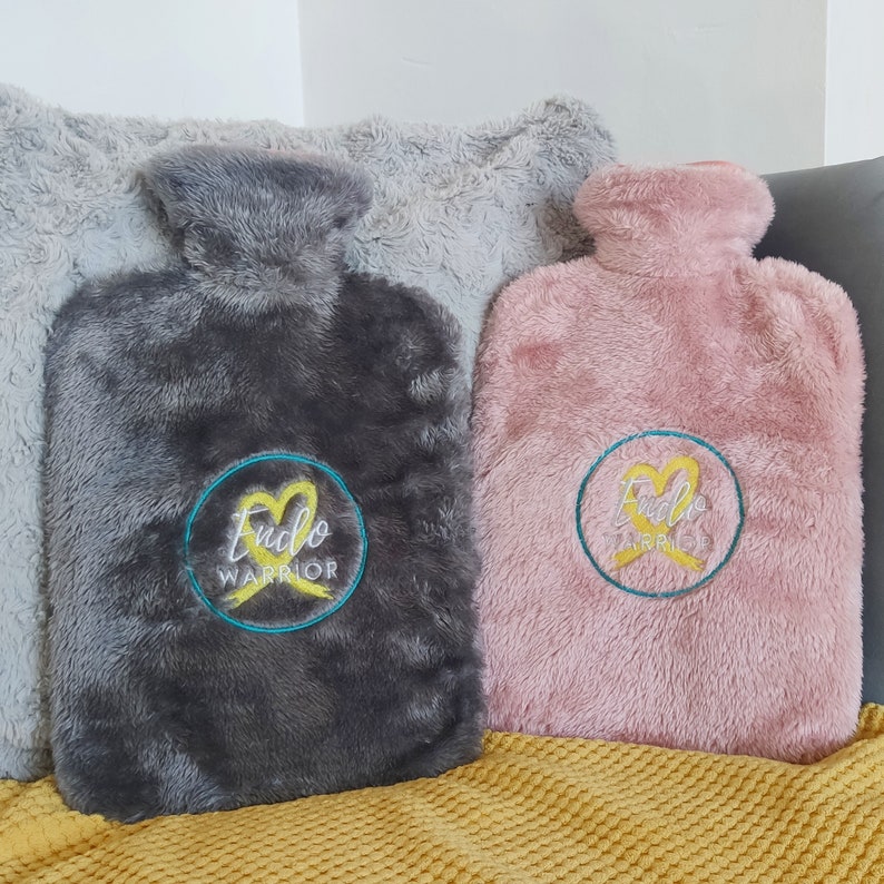 Op de afbeelding: Twee pluche warmwaterkruiken met geborduurde gele linten en de tekst "Endo Warrior" in een cirkel. Een kruik is grijs en de andere is roze.