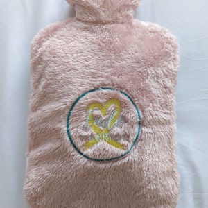 Op de afbeelding: Roze pluche warmwaterkruik met een turquoise cirkel en een geel lint met de tekst "Fido Warrior" geborduurd erop.