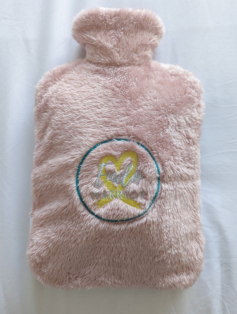 Op de afbeelding: Roze pluche warmwaterkruik met een turquoise cirkel en een geel lint met de tekst "Fido Warrior" geborduurd erop.