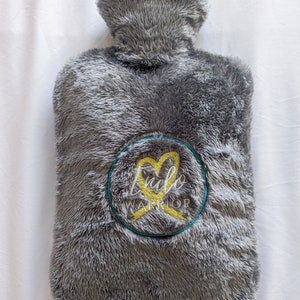 Op de afbeelding: Grijze, pluche warmewaterfles met een geborduurd geel lint en hart. De borduurwerk staat "Endo Warrior".