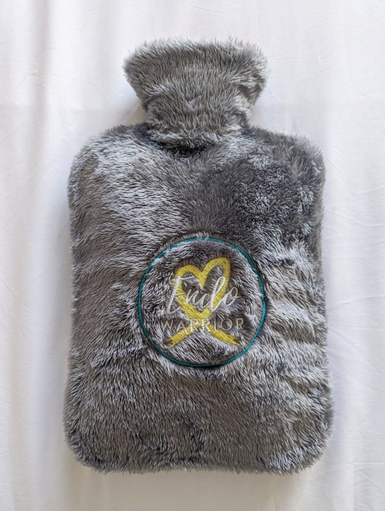 Op de afbeelding: Grijze, pluche warmewaterfles met een geborduurd geel lint en hart. De borduurwerk staat "Endo Warrior".