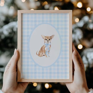 May include: A framed print featuring a watercolor illustration of a Chihuahua dog with a blue bow tie, set against a light blue gingham pattern within an oval. The frame is made of light-colored wood. The background is a blurred Christmas tree.