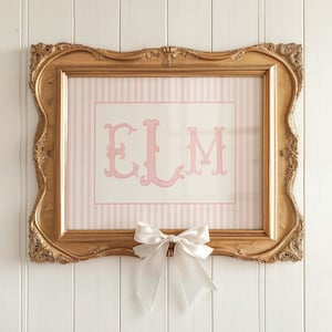 May include: A framed artwork with a gold ornate frame. The artwork features a pink and white striped background with the letters "ELM" in pink. A white bow is attached to the bottom of the frame.