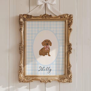 May include: A framed print featuring a brown dachshund wearing a pink and white patterned scarf, set against a light blue gingham background within an oval. The print is in a gold ornate frame, with the name "Milly" below the dog.