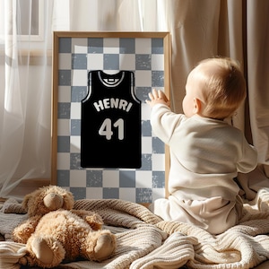 May include: A framed print of a black basketball jersey with the name "HENRI" and the number "41" in white. The background is a blue and white chequered pattern. A teddy bear and a baby are in the foreground.