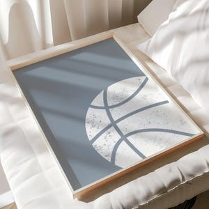 May include: A framed print with a basketball design. The artwork has a light blue background with a distressed white basketball graphic. The frame is a light wood colour, adding a natural touch. This print is suitable for a sports-themed room.