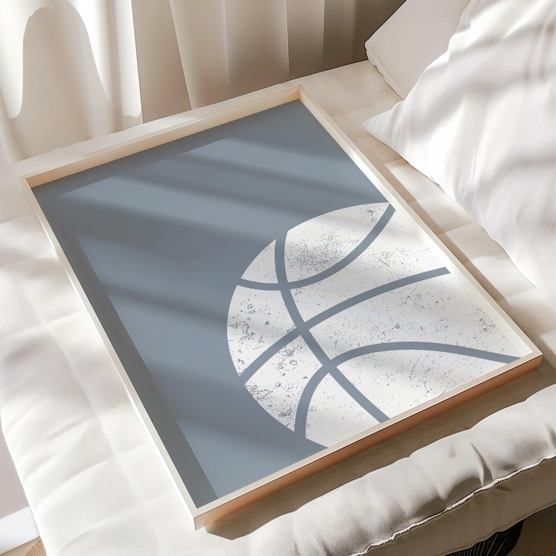 May include: A framed print with a basketball design. The artwork has a light blue background with a distressed white basketball graphic. The frame is a light wood colour, adding a natural touch. This print is suitable for a sports-themed room.
