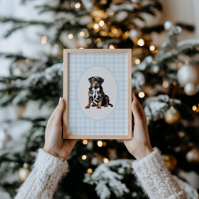 May include: A framed print featuring a Rottweiler puppy with a blue bow tie, set against a light blue gingham background. The print is in a light wood frame. The background is a blurred Christmas tree with lights.