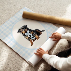 May include: A watercolor print of a Rottweiler puppy wearing a blue bow, set against a light blue gingham pattern. The artwork is on a large, rolled-up poster, with a second rolled poster in the background.