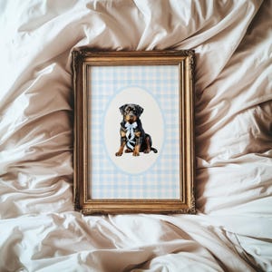 May include: A framed print featuring a Rottweiler dog with a blue bow. The dog is centered in an oval against a light blue and white checkered background. The frame is gold-colored and ornate.