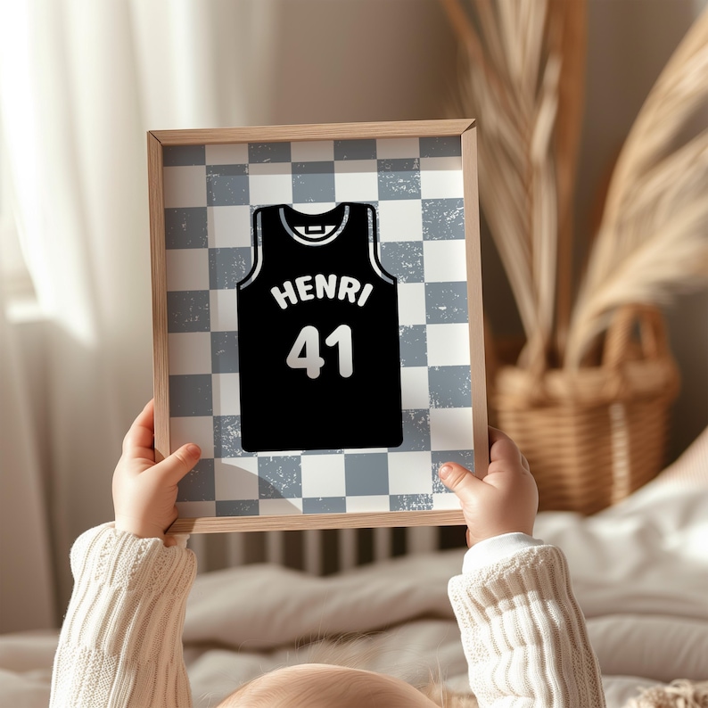 May include: A framed print showing a black basketball jersey design with the name "HENRI" and the number "41" in white. The background has a grey and white checkered pattern. The frame is light-coloured wood.