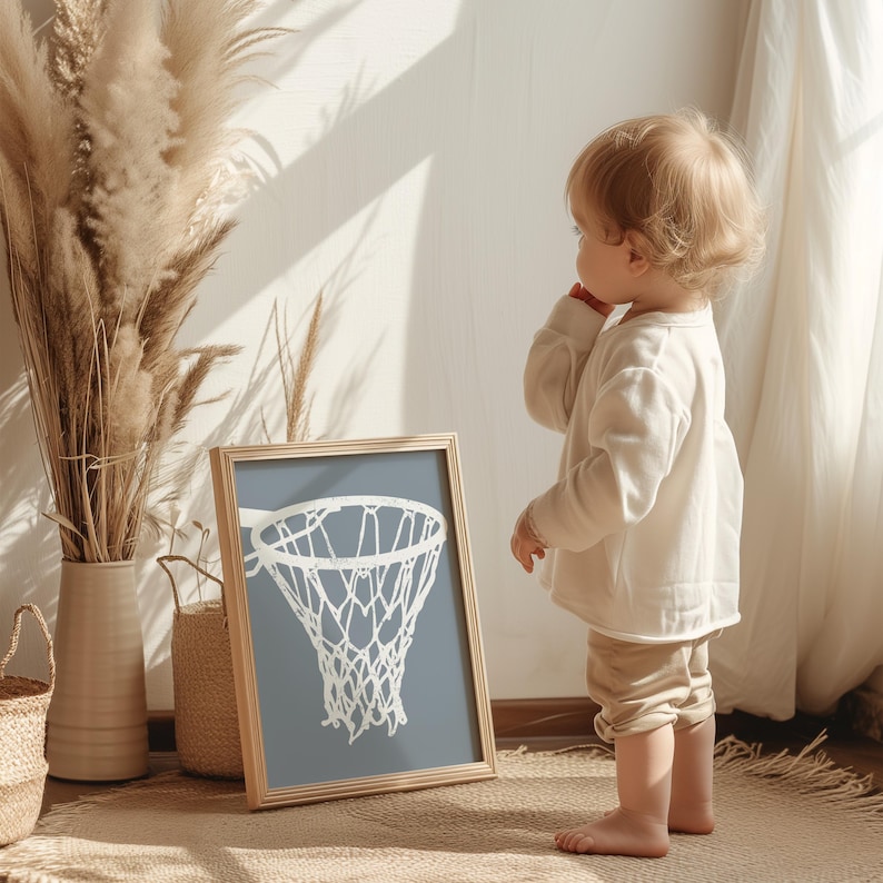 May include: A framed print of a basketball hoop with a white net against a blue-grey background. The wooden frame sits on a rug, next to a vase of dried plants. A child in a cream-coloured outfit stands nearby.