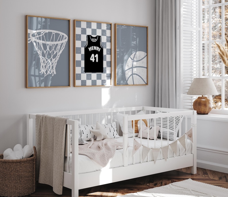 May include: Three framed basketball-themed prints in a nursery. One shows a basketball net, another a black jersey with "Henri 41", and the third a basketball. The prints are above a white cot with decorative pillows and a blanket.