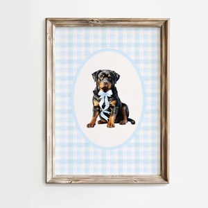 May include: A framed print featuring a Rottweiler dog with a blue bow tie, set against a light blue and white gingham pattern. The dog is centered within a light blue oval, adding a touch of elegance to the artwork. The frame is a natural wood color.