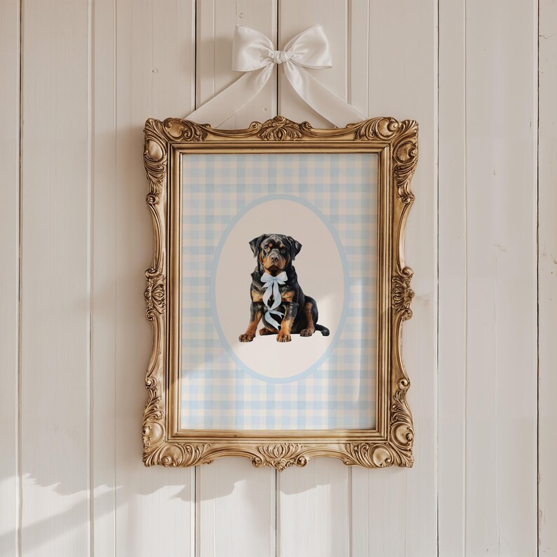 May include: A framed print featuring a Rottweiler puppy with a blue bow, set against a light blue and white gingham background within an oval. The ornate gold frame is topped with a white bow and ribbon, hanging on a white wooden wall.