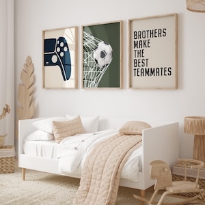 Op de afbeelding: Drie ingelijste prints met een gamecontroller, een voetbal in een net en de tekst "BROTHERS MAKE THE BEST TEAMMATES". De prints bevinden zich boven een wit bed met een beige dekbed en kussens.