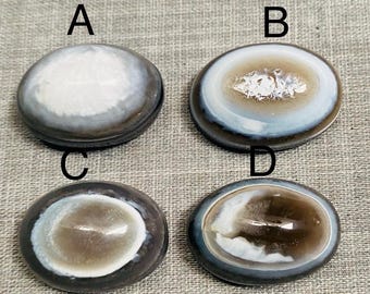 May include: Four oval stones with a polished finish. Each stone has a lighter central area and a darker, banded border. The stones are labelled A, B, C, and D. The stones are displayed on a grey surface.