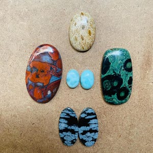 May include: Assortment of six oval-shaped cabochon gemstones. The stones are in various colors and patterns, including red, brown, white, blue, green, and black.