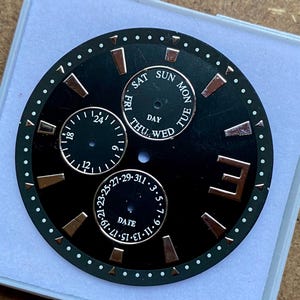 Puede incluir: Una esfera de reloj negra con detalles dorados y puntos blancos alrededor del perímetro. La esfera muestra el día, la fecha y la hora, con subesferas para cada uno. El texto incluye los días de la semana y números.