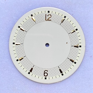 Pode incluir: Um mostrador de relógio vintage com fundo creme e um padrão geométrico decorativo. Possui marcadores de hora pretos e dourados, com os números 12 e 6 em dourado. Um pequeno orifício está centrado no mostrador.