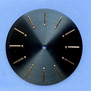 Puede incluir: Una esfera de reloj negra con marcadores de hora dorados y un agujero central. Los marcadores de hora son rectangulares con un pequeño punto al final. La esfera tiene una textura de metal cepillado.