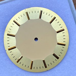 Pode incluir: Um mostrador de relógio dourado com uma abertura circular central e uma série de pequenos orifícios ao redor do perímetro. O mostrador apresenta marcadores de hora e minuto retangulares dourados. O design geral é simples e elegante.