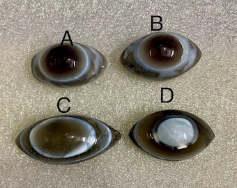 May include: Four oval gemstone cabochons, each resembling an eye. The stones feature concentric circles of brown, white, and grey, with a glossy finish. The cabochons are labelled A, B, C, and D.