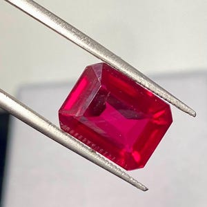 May include: A vibrant, deep red gemstone with an emerald cut, featuring sharp, clean facets that reflect light. The rectangular stone is held securely by a pair of silver-toned metal tweezers, showcasing its intense color and transparent clarity.