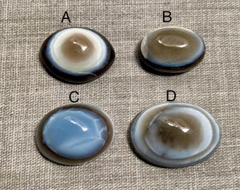 May include: Four oval cabochons with eye-like patterns. The cabochons are in shades of brown, blue, and white, with a glossy finish. They are arranged on a grey textured surface. Suitable for jewellery making or other crafts.