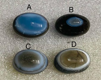 May include: Four polished, oval gemstones in a variety of colours. Gem A is blue, Gem B is black with a blue centre, Gem C is grey and white, and Gem D is brown and white. Each stone has a dark border.